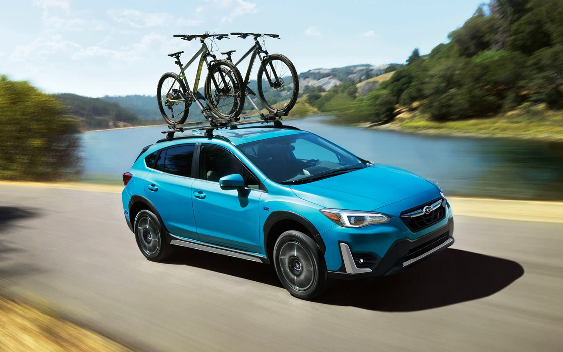 A blue Crosstrek Hybrid with two bicycles on its roof rack driving beside a river | Open Road Subaru in Union NJ